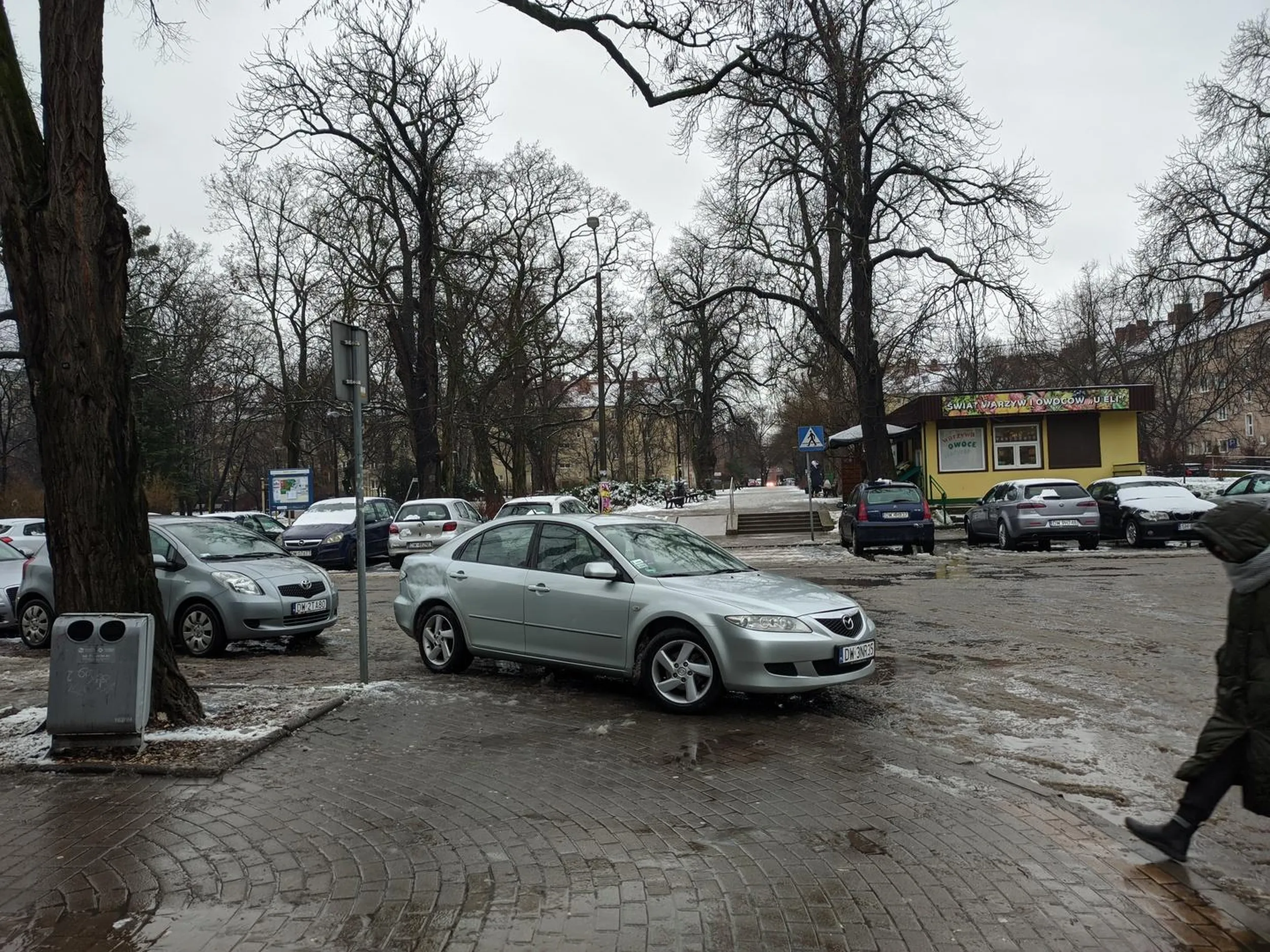 DW3NR35 - samochód marki Mazda zaparkowany na środku przejścia dla pieszych. Ludzie wychodzą na jezdnię za pasami. Kierowcy jadący ulicą mają utrudnioną widoczność. Starsi ludzie nie mają problemy z obejściem samochodu.