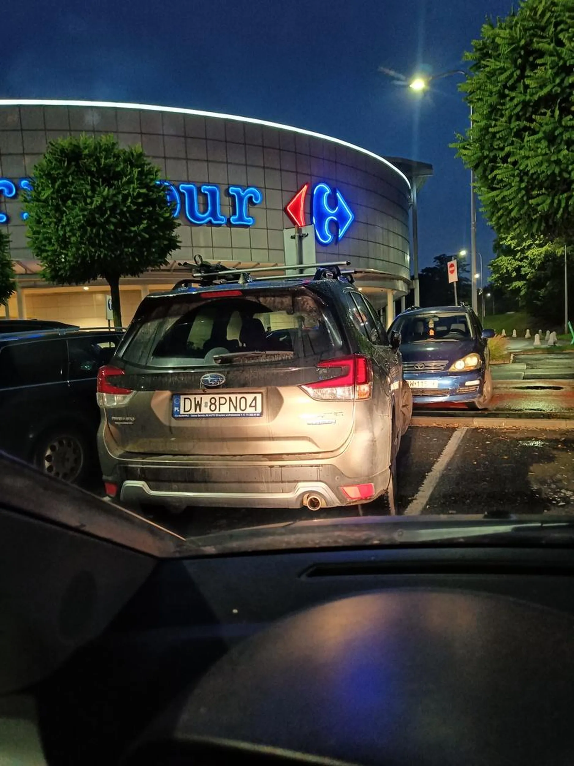 Parkuje pod kątem, część jego samochodu wystaje na sąsiednie miejsce parkingowe, co utrudnia wyjazd innym kierowcom.
