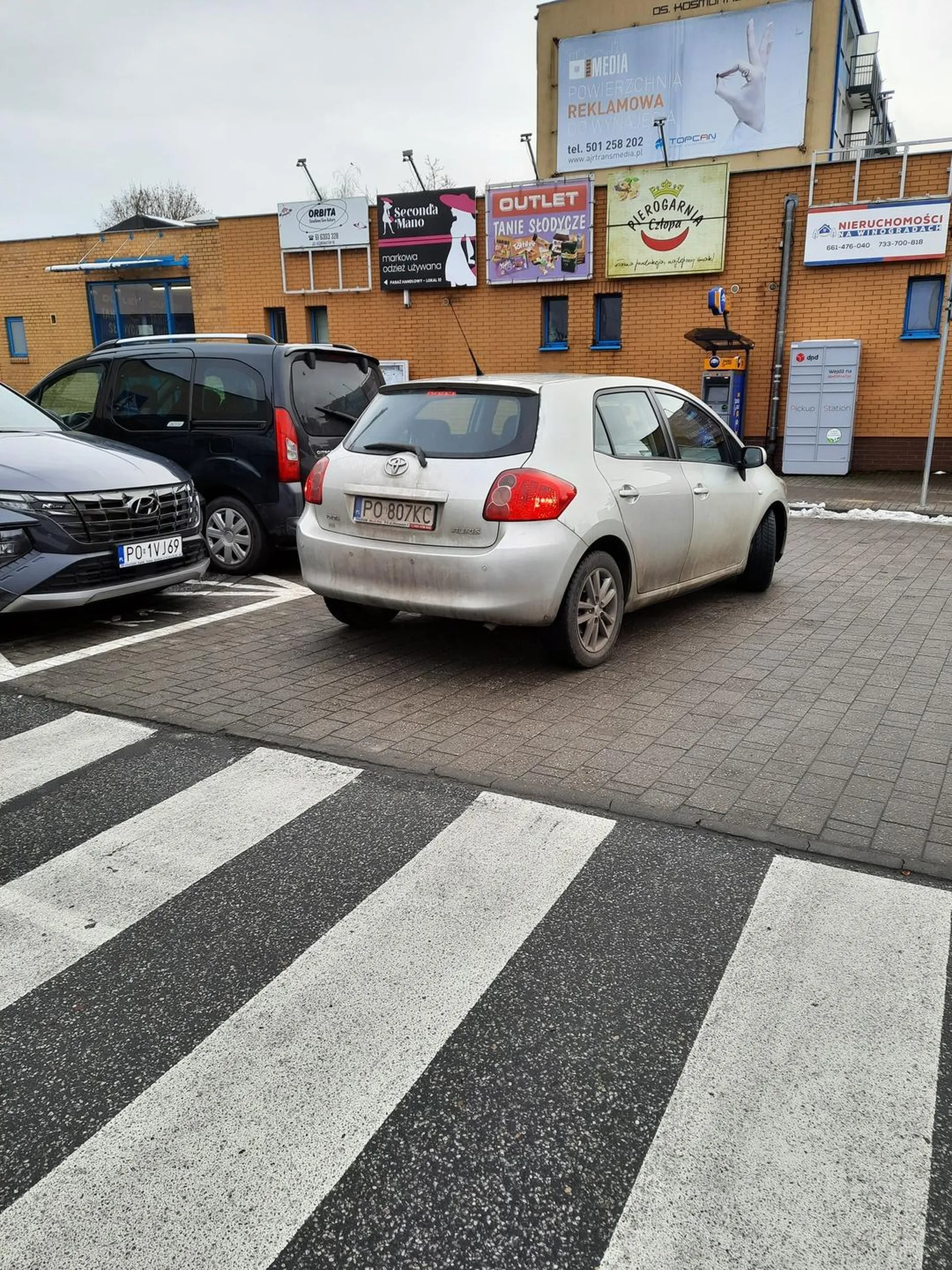 Pani rozmawi przez telefon stoi po lewej stronie blokując 1 pas ruchu wjazdu na parking.