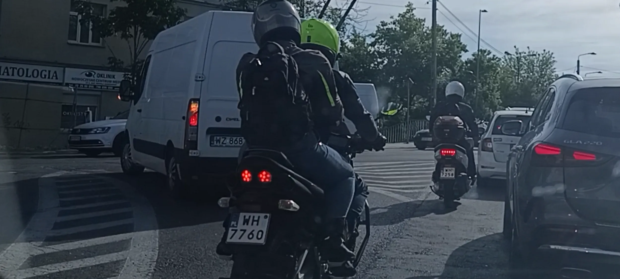 Bandyta na wraku skutera przepycha się w korku między autami, wyprzedza na podwójnej linii ciągłej przed przejściem dla pieszych i na samym przejsciu dla pieszych.