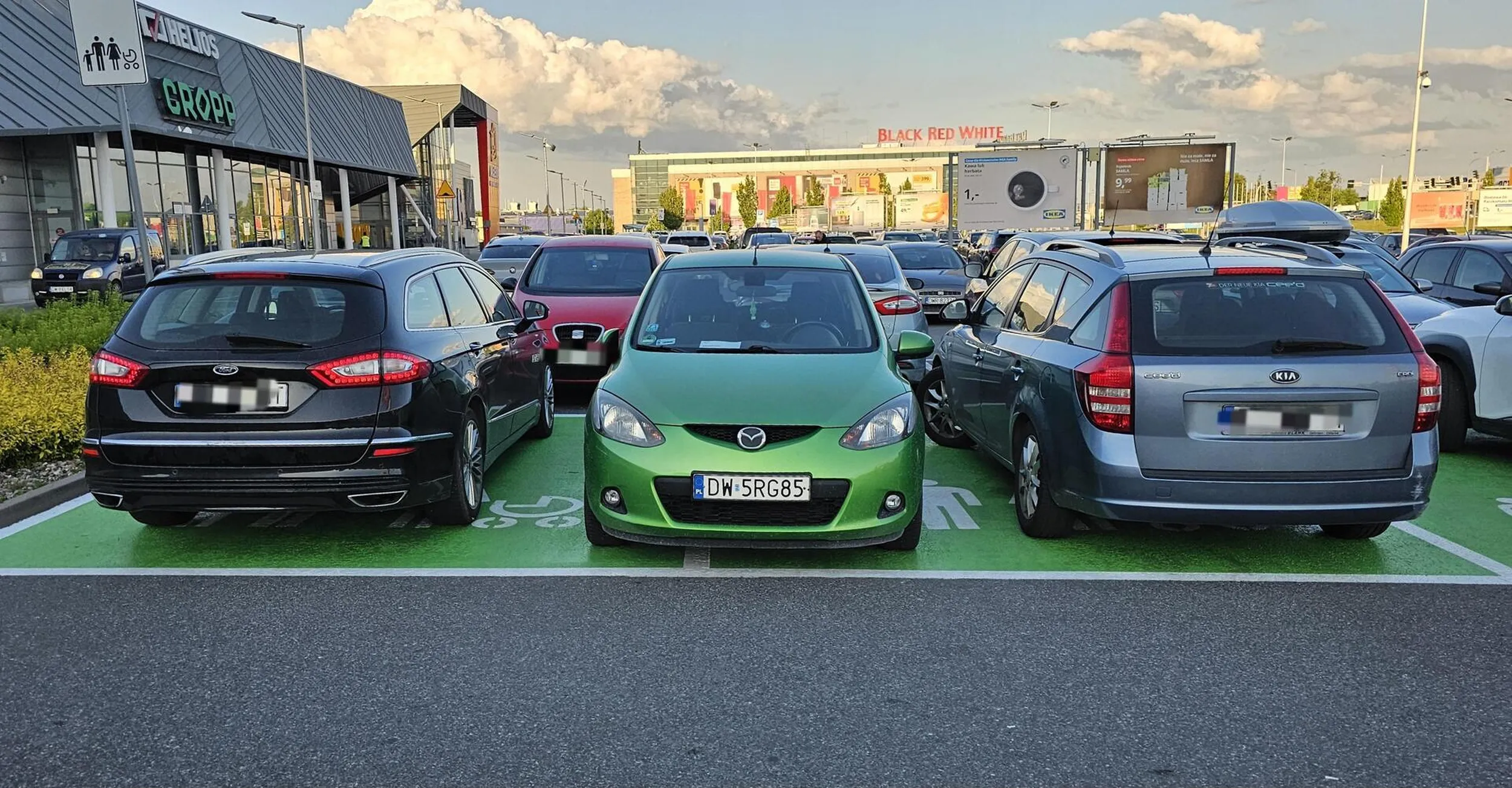 Kierowca zielonej Mazdy zaparkował skrajnie egoistycznie, centralnie na linii, zabierając przestrzeń sąsiednim miejscom. Swoim brakiem wyobraźni i elementarnej kultury uniemożliwił samotnemu rodzicowi bezpieczne wsadzenie dziecka do auta. To klasyczny przykład parkingowego chamstwa: zero empatii, zero odpowiedzialności, pełne skupienie wyłącznie na własnej wygodzie kosztem innych.