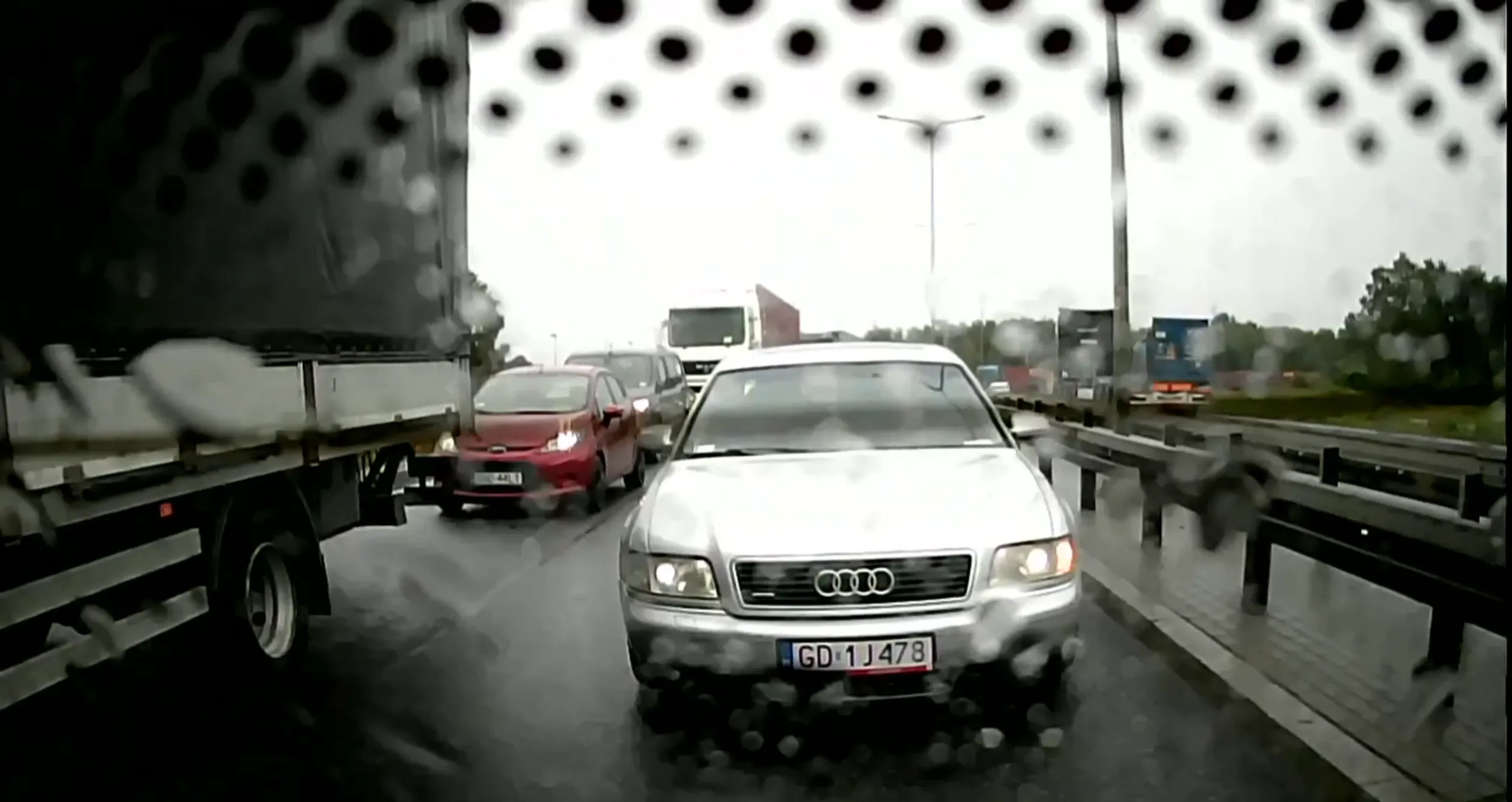 Przechlany patus, jedzie na zderzaku w czasie deszczu, pokazuje środkowy palec i do tego pedau