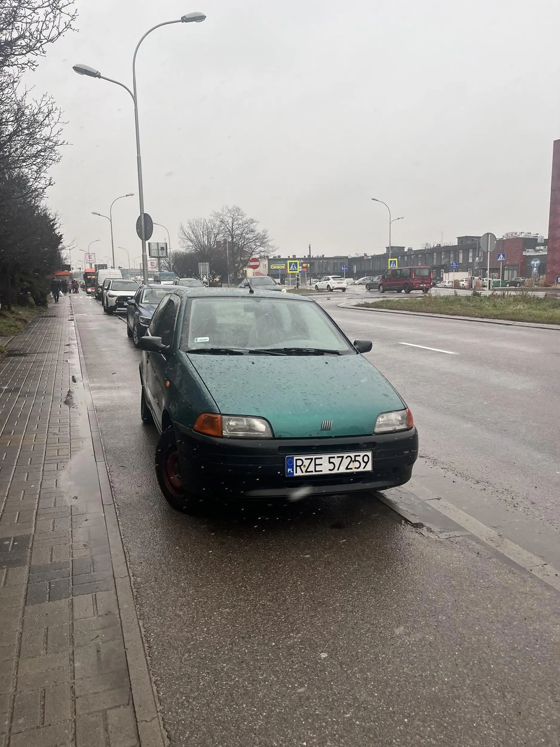 Na zdjęciu widać samochód zaparkowany na drodze rowerowej, co jest niezgodne z przepisami ruchu drogowego w Polsce. Droga rowerowa jest przeznaczona wyłącznie dla ruchu rowerzystów i nie wolno na niej zatrzymywać się ani parkować pojazdów, chyba że przepisy ruchu drogowego (lub oznakowanie) wyraźnie to dopuszczają, czego w tym przypadku nie widać. Art. 49 ust. 1 pkt 11 ustawy Prawo o ruchu drogowym stanowi, że zabronione jest zatrzymywanie pojazdu “na drogach dla rowerów”. Za takie parkowanie kierowca może otrzymać mandat karny, a w skrajnych przypadkach pojazd może zostać odholowany. Dodatkowo, parkowanie w ten sposób stwarza zagrożenie i utrudnia ruch rowerzystom, co może być szczególnie niebezpieczne w przypadku słabej widoczności, np. podczas opadów, jak na zdjęciu.